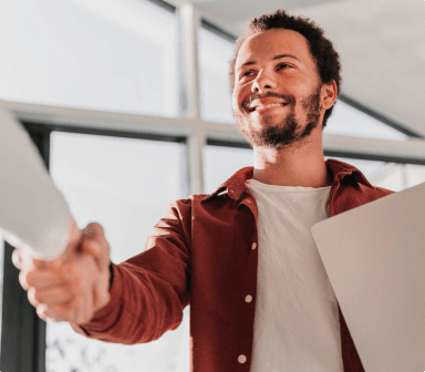 Na imagem vemos um homem segurando um envelope e sorrindo enquanto aperta a mão de outra pessoa, o homem veste uma blusa vermelha e com uma camisa branca por baixo