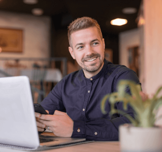 Imagem de um homem sentado em uma mesa com um notebook a sua frente segurando um celular e sorrindo, o homem veste uma camisa social azul escuro