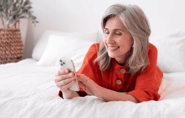 Imagem de uma mulher deitada na cama sorrindo enquanto utiliza um telefone celular, a mulher veste camisa vermelha