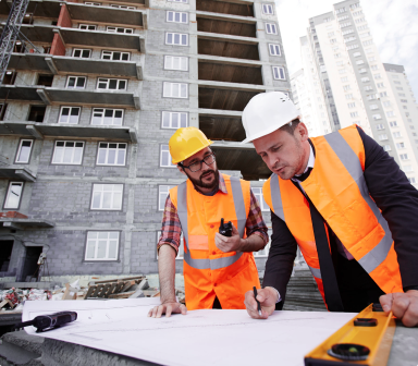 Imagem de dois engenheiros conversando sobre um edifício, um deles está com a planta aberta sobre uma mesa